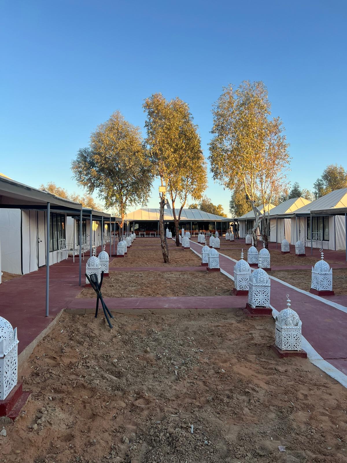Vue extérieure d'un campement désertique avec une allée pavée en rouge, des lanternes décoratives blanches de chaque côté et des bâtiments/tentes sous un ciel bleu clair.