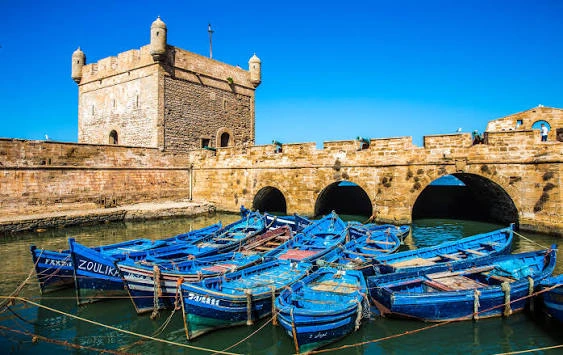Barques de pêche bleues amarrées dans le port d'Essaouira avec la tour de la Sqala du Port et des ponts en pierre sous un ciel bleu clair.