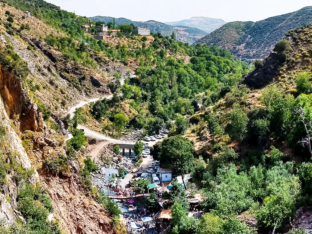 Vue aérienne de la vallée de l'Ourika, au Maroc, avec des collines rocheuses et verdoyantes encadrant une rivière en bas. Une route sinueuse mène à une zone d'activités avec des parking et des installations en bord de l'eau. Au sommet d'une colline, on aperçoit des bâtiments traditionnels.