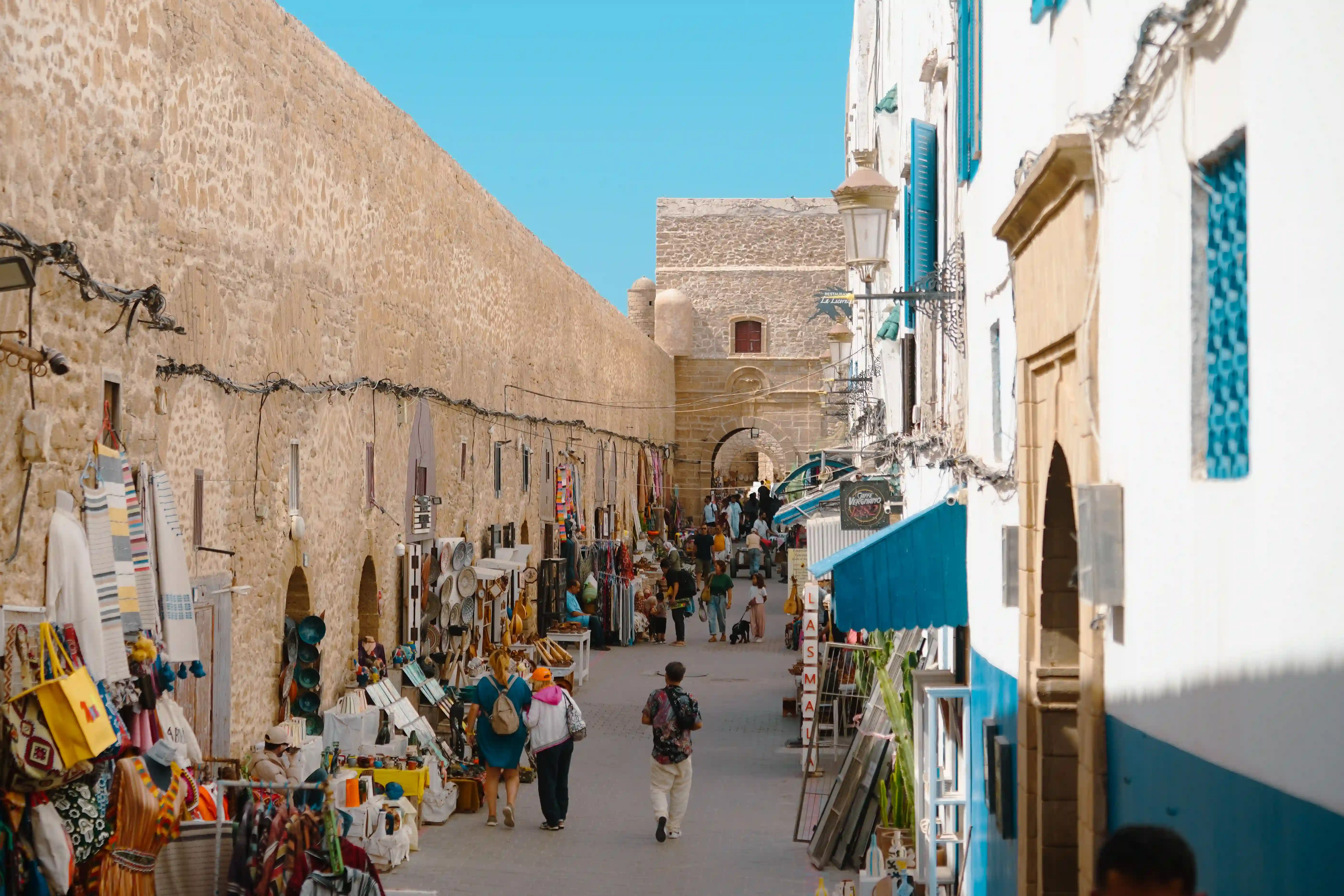 Vue aérienne d'une rue animée de la médina d'Essaouira, flanquée d'un mur d'enceinte en pierre d'un côté et de bâtiments blancs aux accents bleus de l'autre. Des boutiques locales et des stands exposent de l'artisanat,
