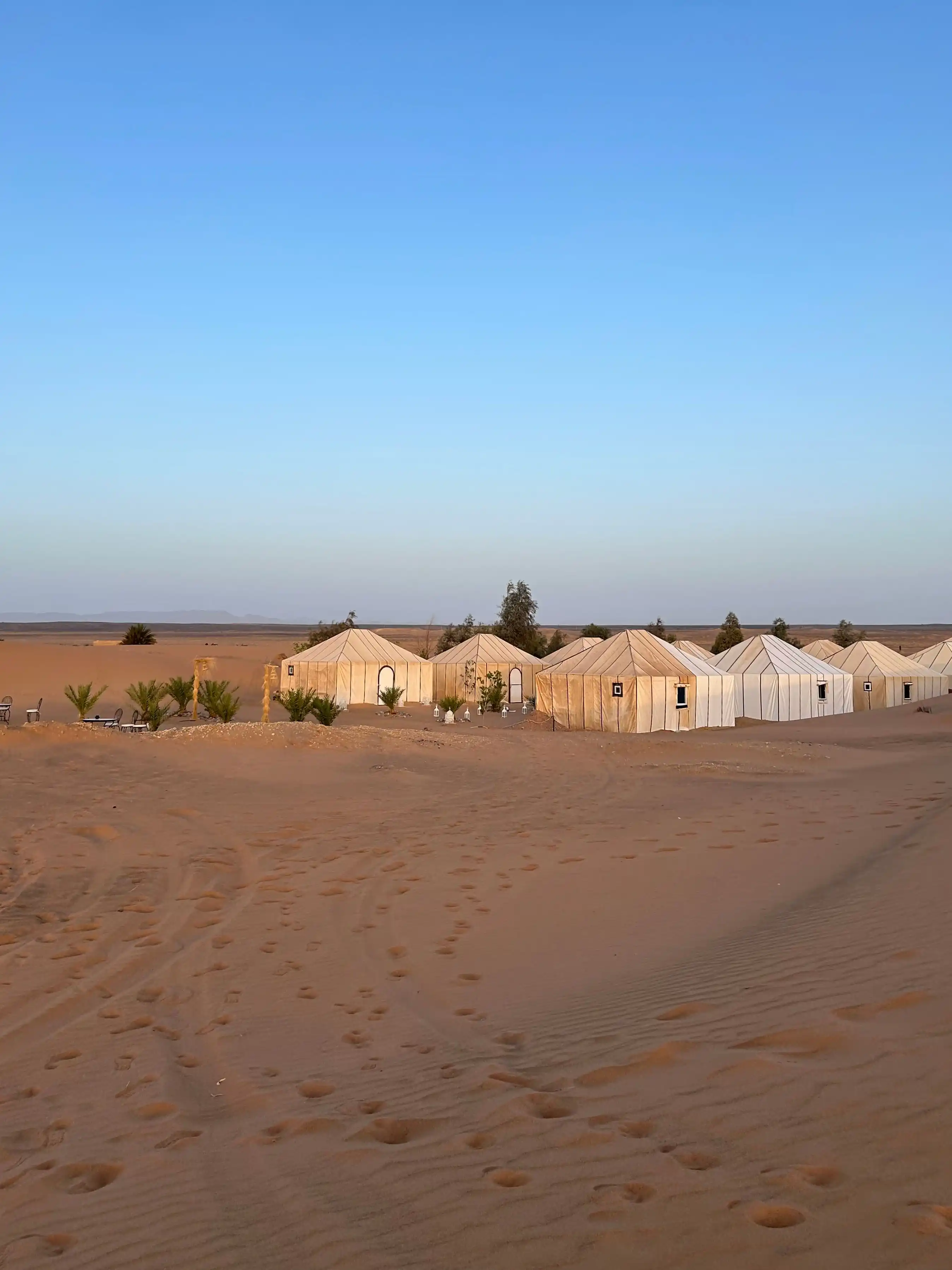 Campement de luxe composé de tentes blanches installées au cœur des dunes de sable du désert sous un ciel bleu dégagé.